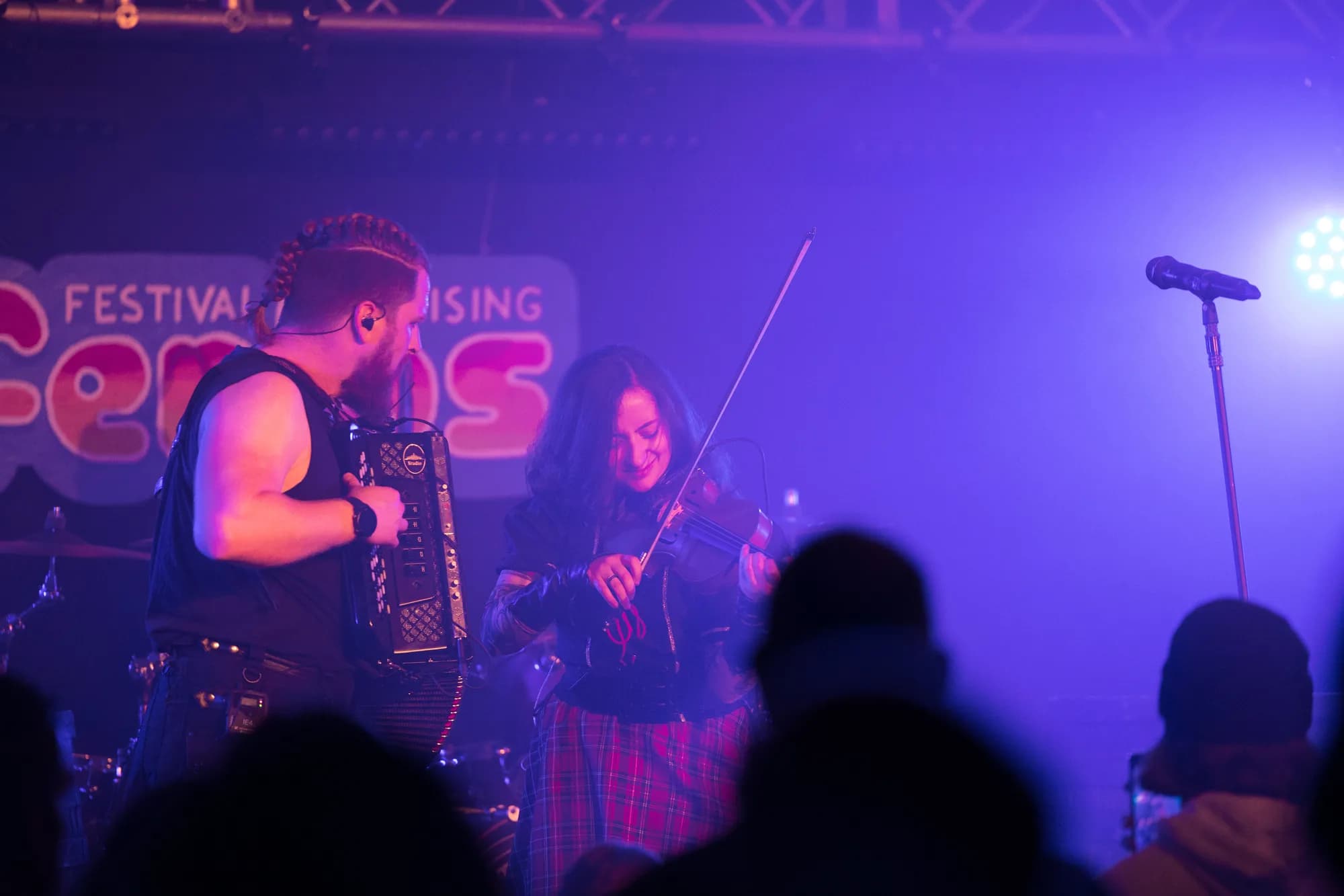 A male accordion player and female violinist perform on a stage bathed in purple light with a festival banner behind them.
