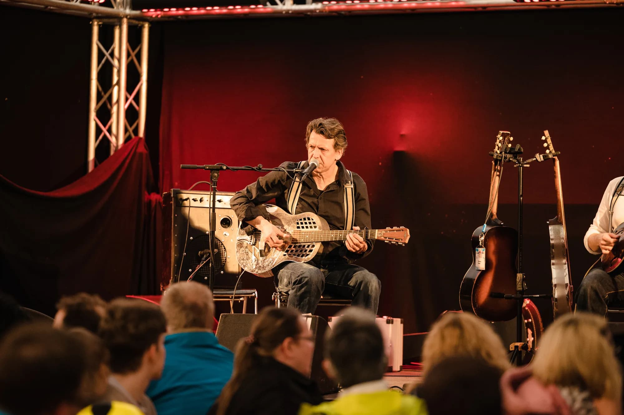 A male musician with a resonator guitar sings into a microphone on a stage with red lighting.