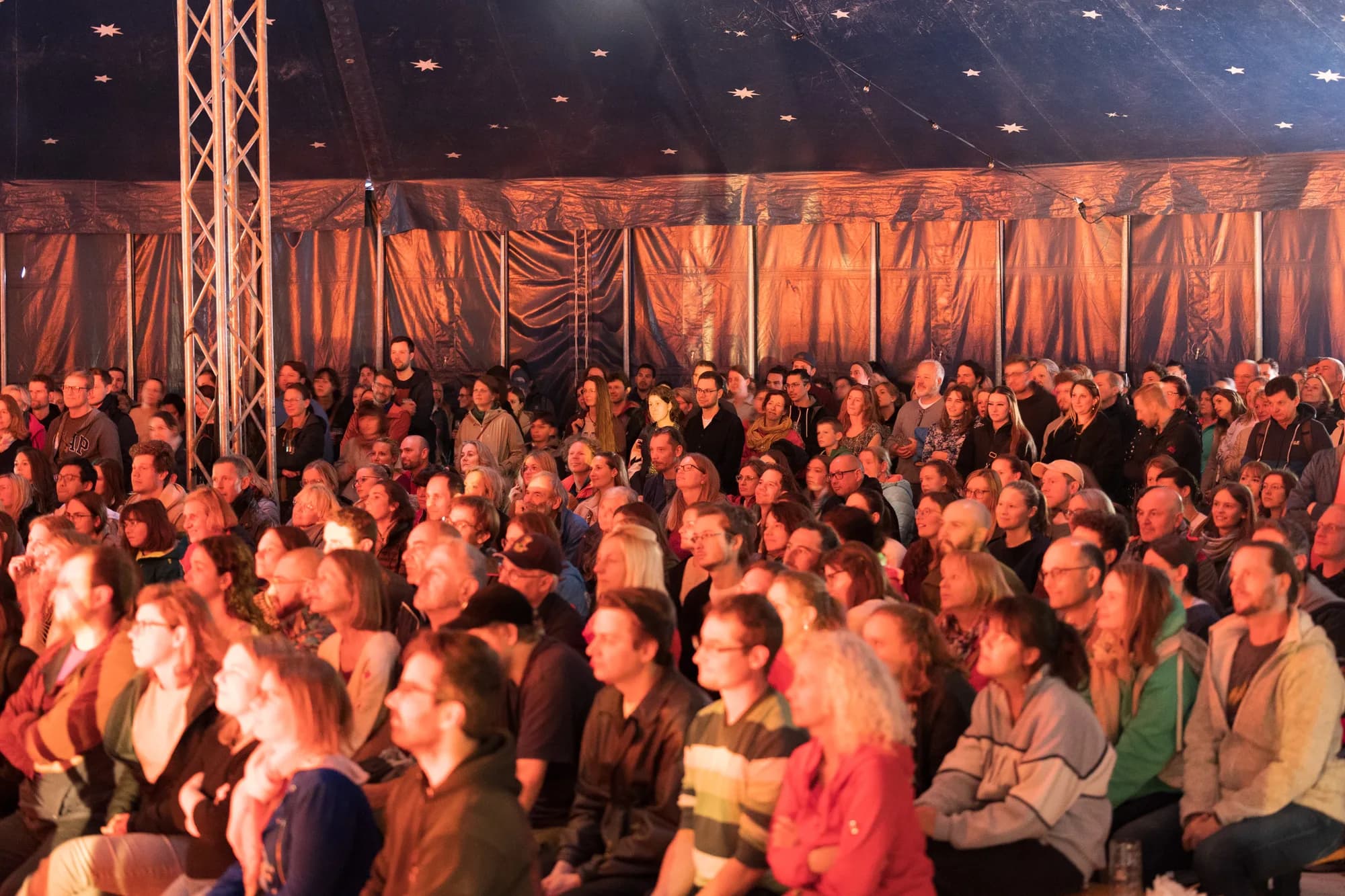 A large crowd of diverse festival-goers sits closely together inside a dimly lit tent, watching a performance.