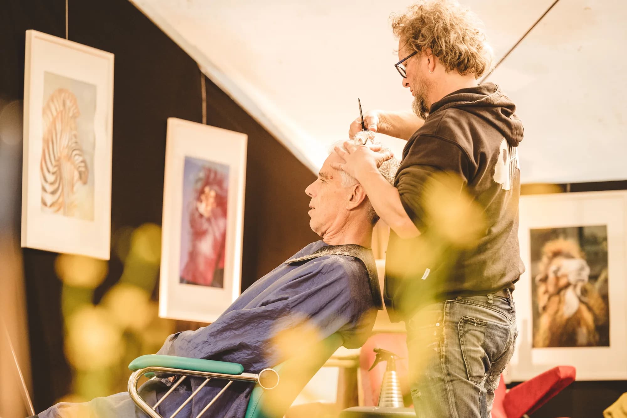 A barber with curly hair gives a haircut to an older man in a blue cape inside a tent with art on the walls.