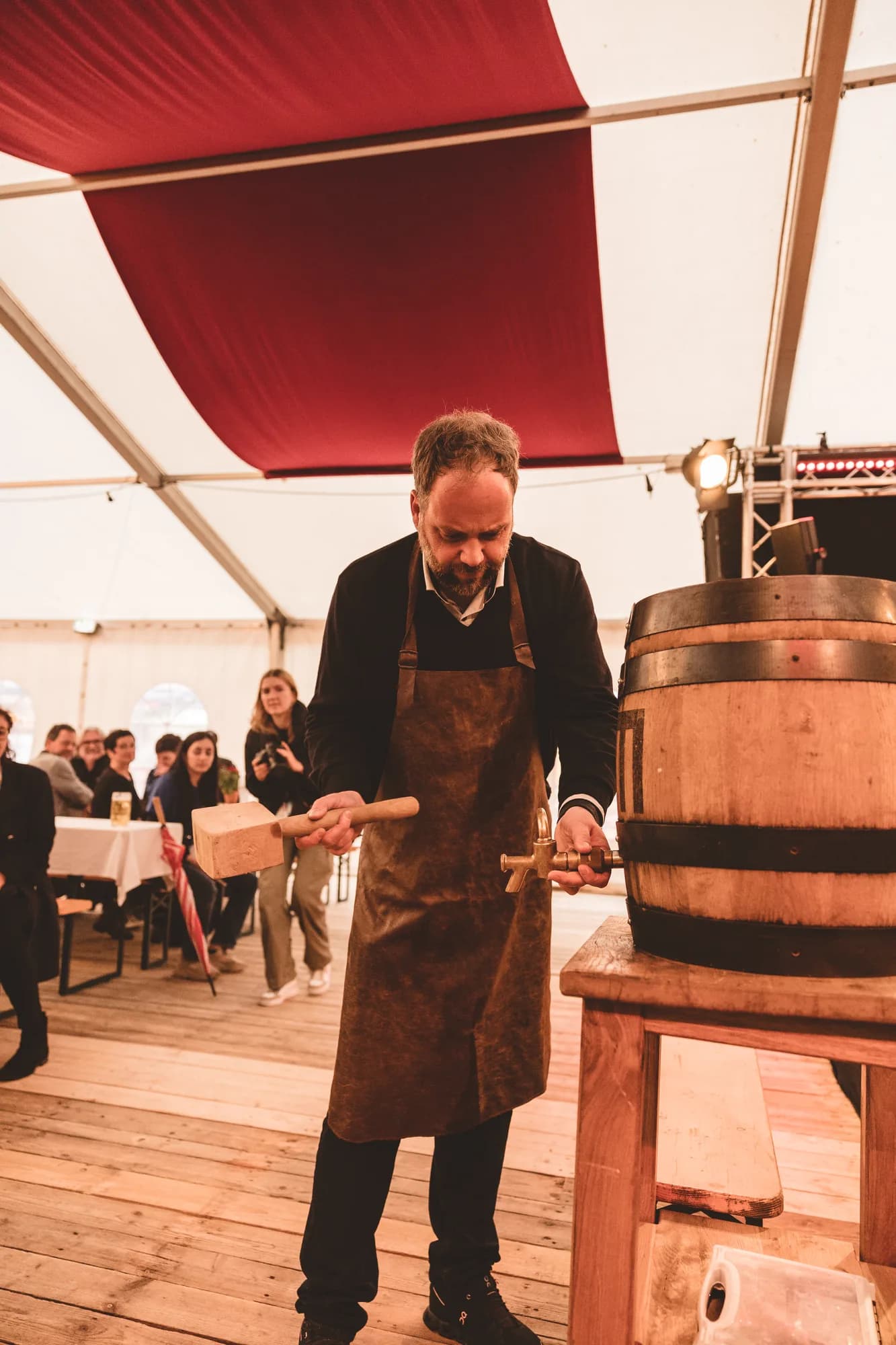 A man in a leather apron taps a wooden barrel with a mallet inside a festive white tent.