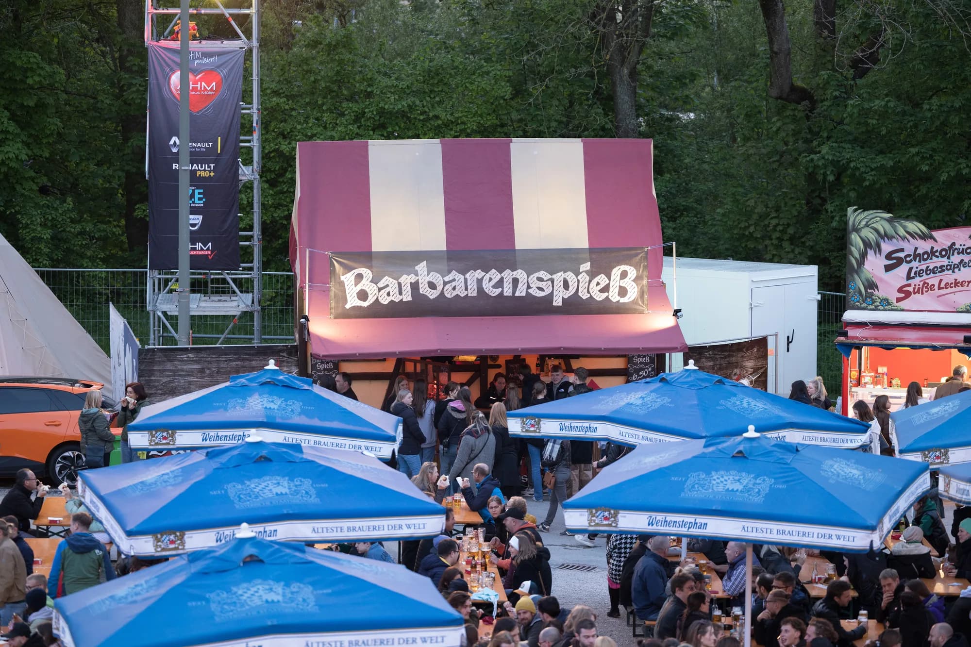 A busy beer garden scene at the Uferlos Festival featuring a food stall named Barbarenspieß and rows of blue beer umbrellas.