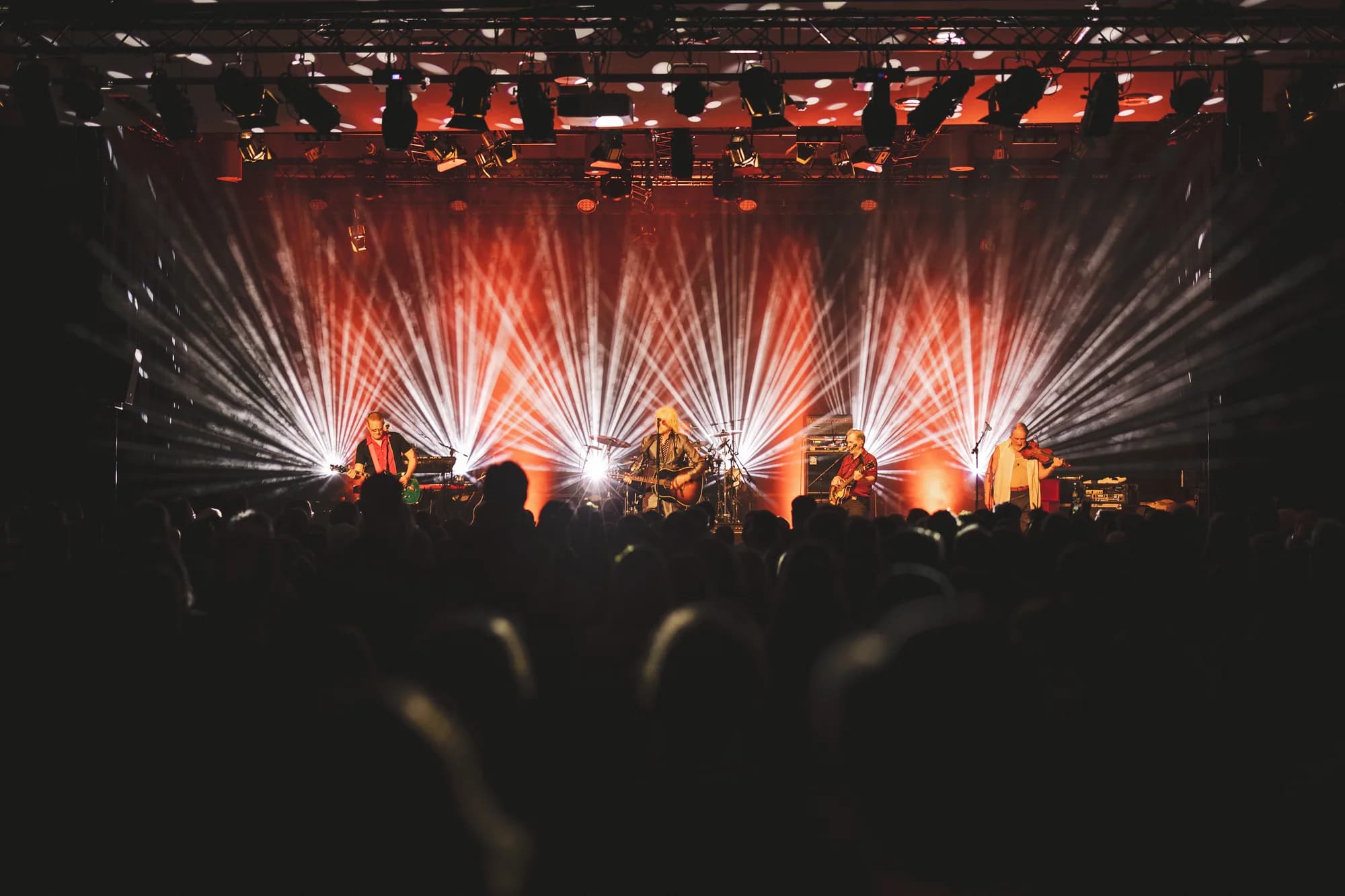 A brass band performs on stage bathed in dramatic orange and white spotlights while a crowd watches in the dark.