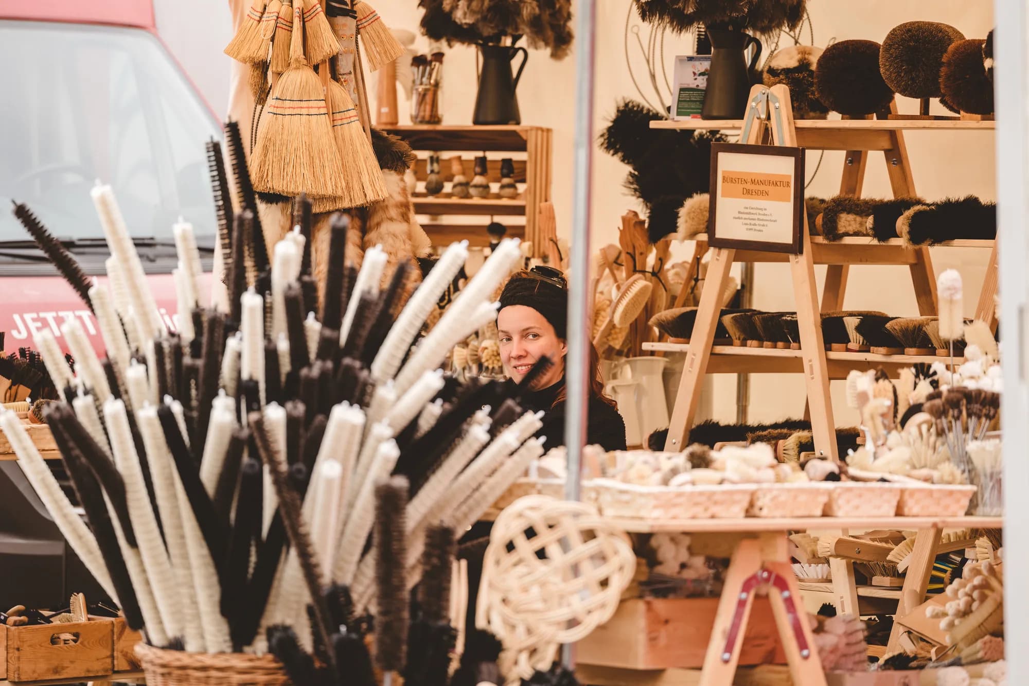 A vendor smiles from behind a rustic stall filled with handmade brooms and brushes.