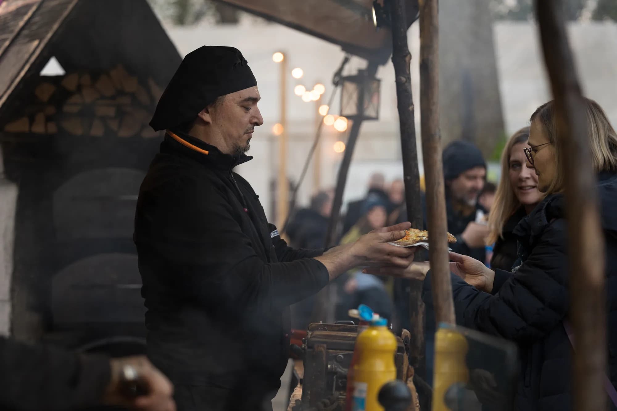 A chef in a black uniform hands a slice of pizza to a smiling customer at an outdoor food stall.