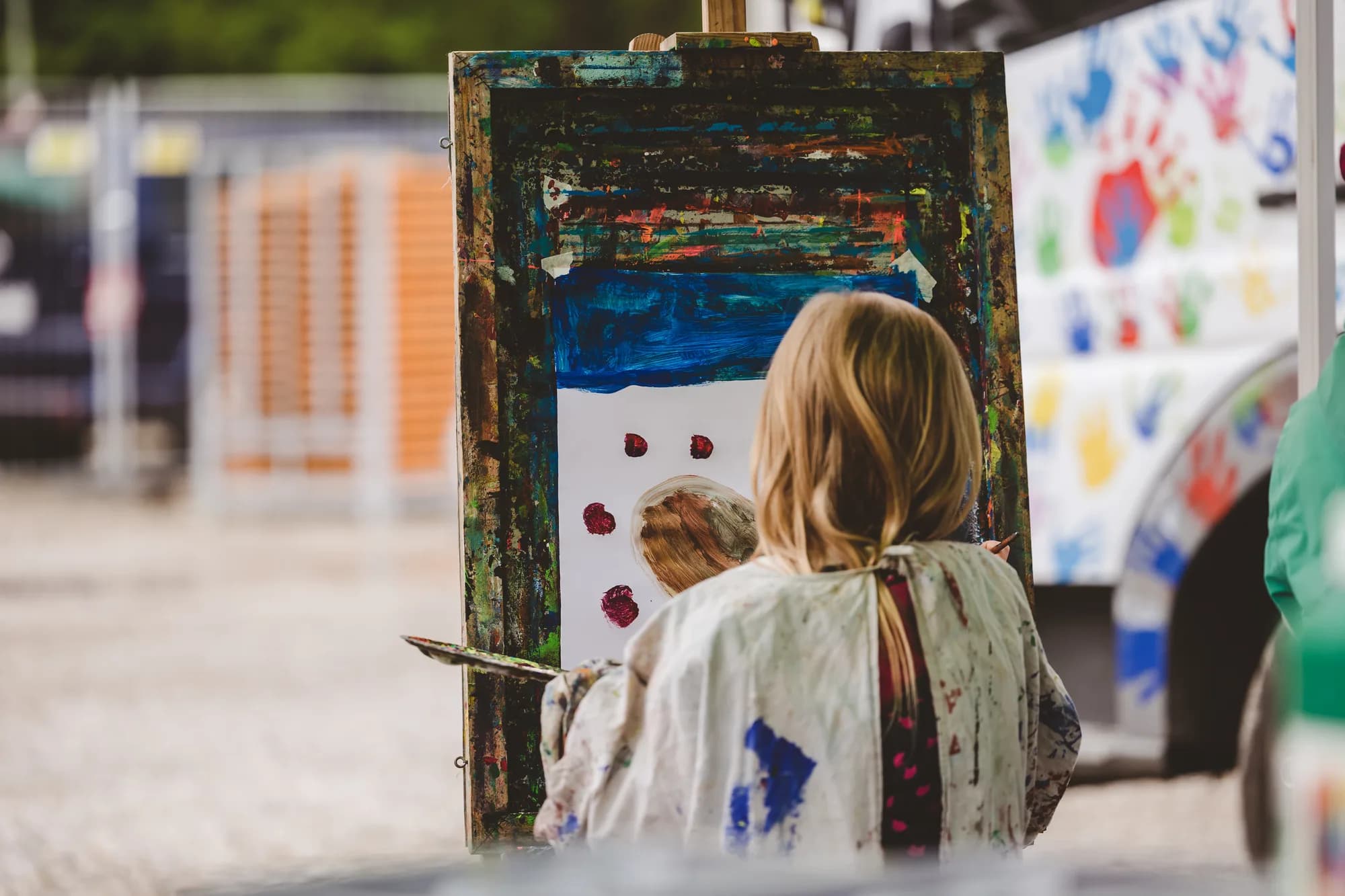 A young girl with blonde hair wearing a paint-splattered smock paints on an easel at the children's tent.