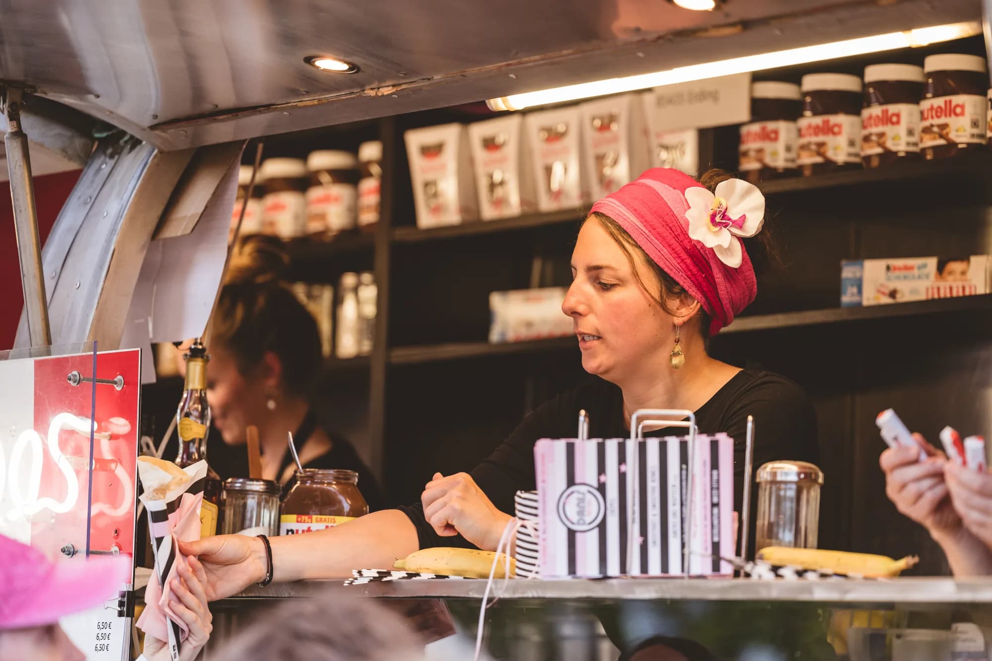 A female vendor wearing a pink headscarf serves a crepe to a customer at a festival food stall.