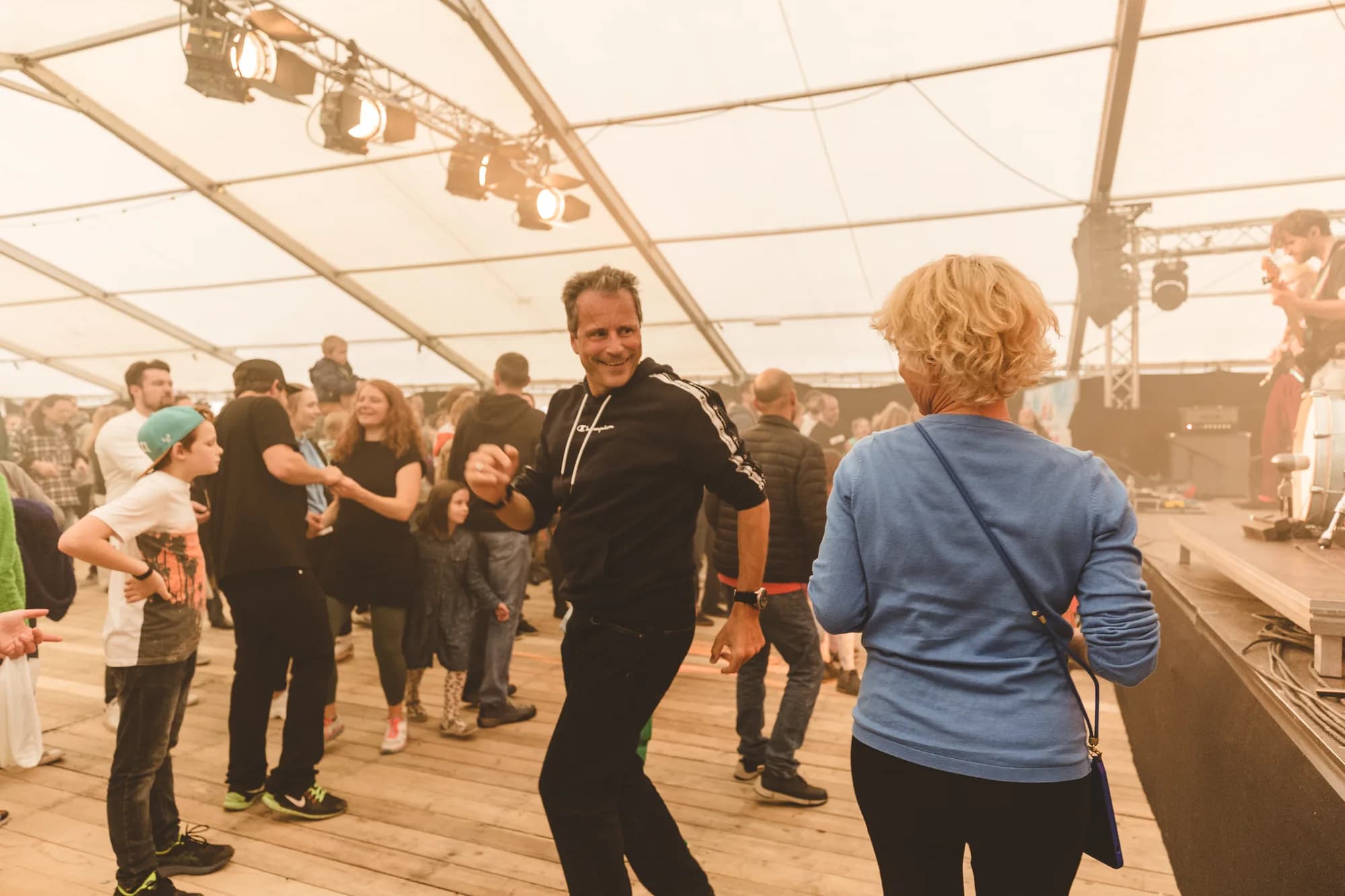 A smiling man in a black tracksuit dances energetically on a wooden stage floor while a DJ performs in the background.