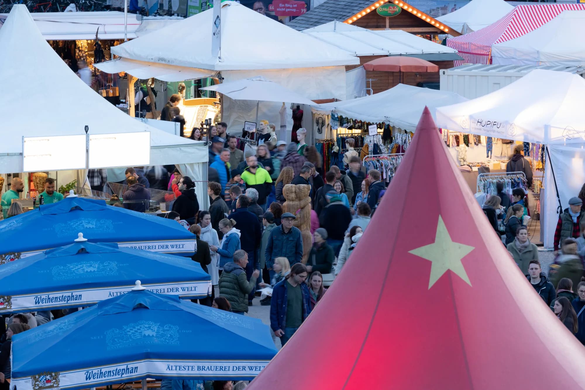 A dense crowd of festival-goers walks between white vendor tents and blue umbrellas during the evening.