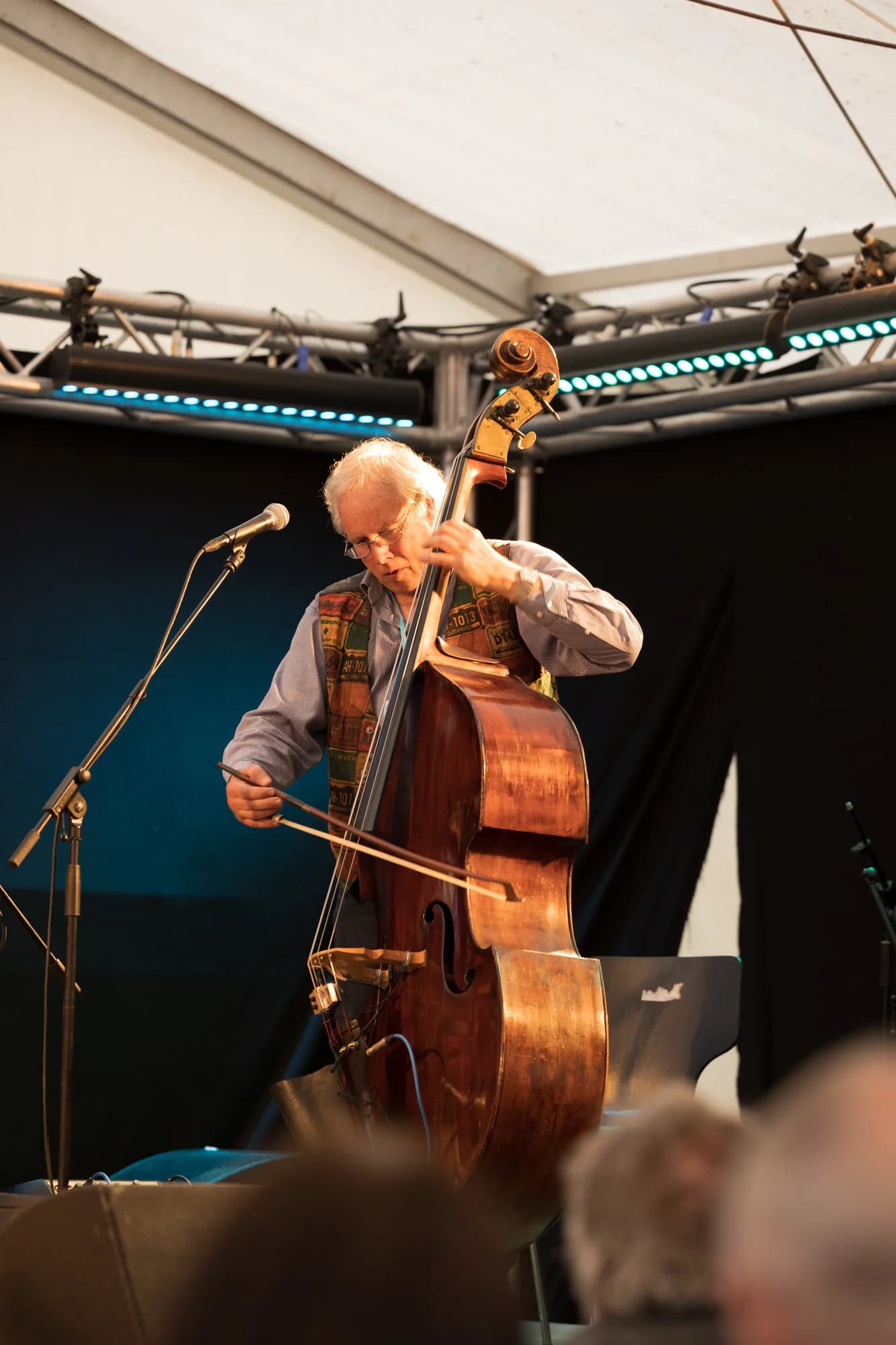 An older musician with white hair plays a wooden double bass with a bow on a festival stage.