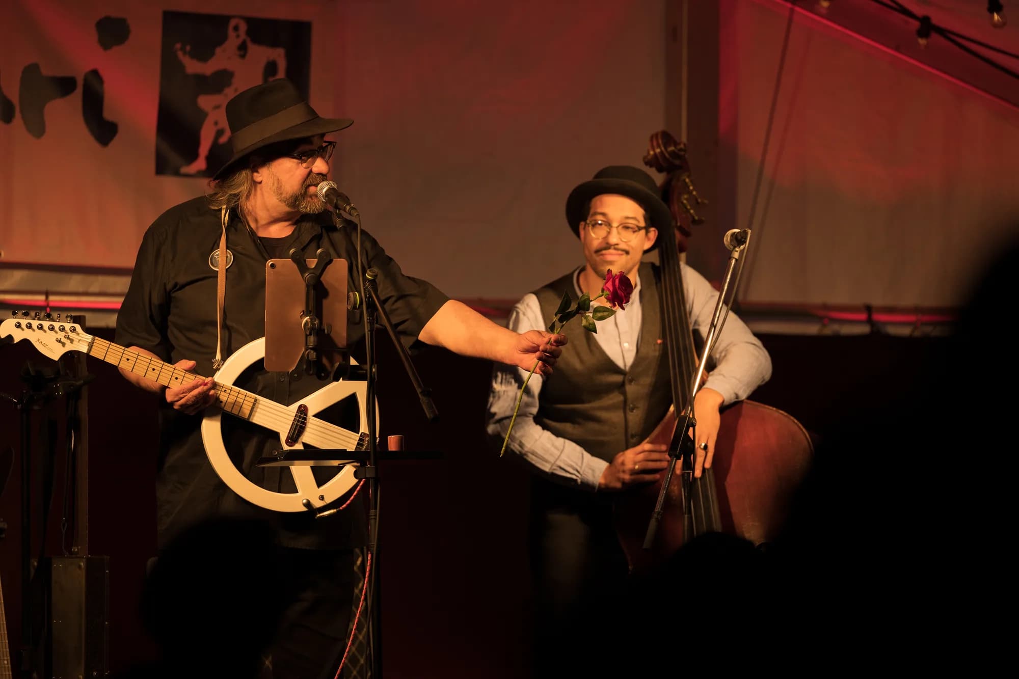 A bearded guitarist in a hat sings into a microphone while offering a red rose to a cellist wearing a vest and glasses on a dimly lit stage.