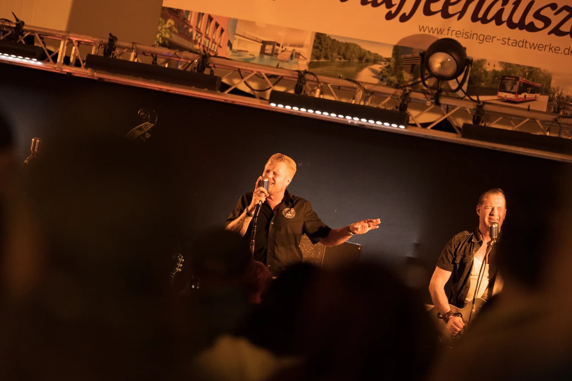 Two male singers perform on stage at night, illuminated by warm spotlights with a banner for Freisinger Stadtwerke visible above them.