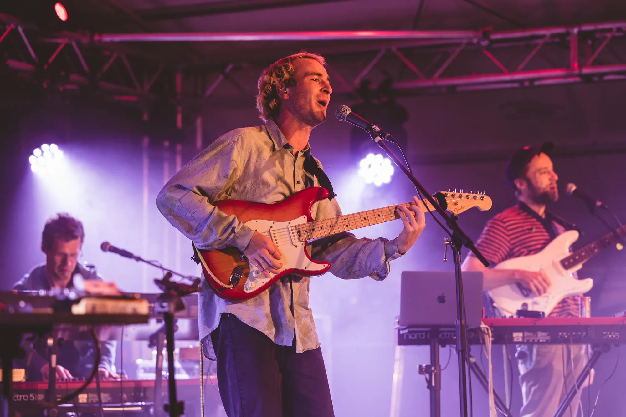 A male musician with curly hair sings and plays a red electric guitar on a stage bathed in purple light.