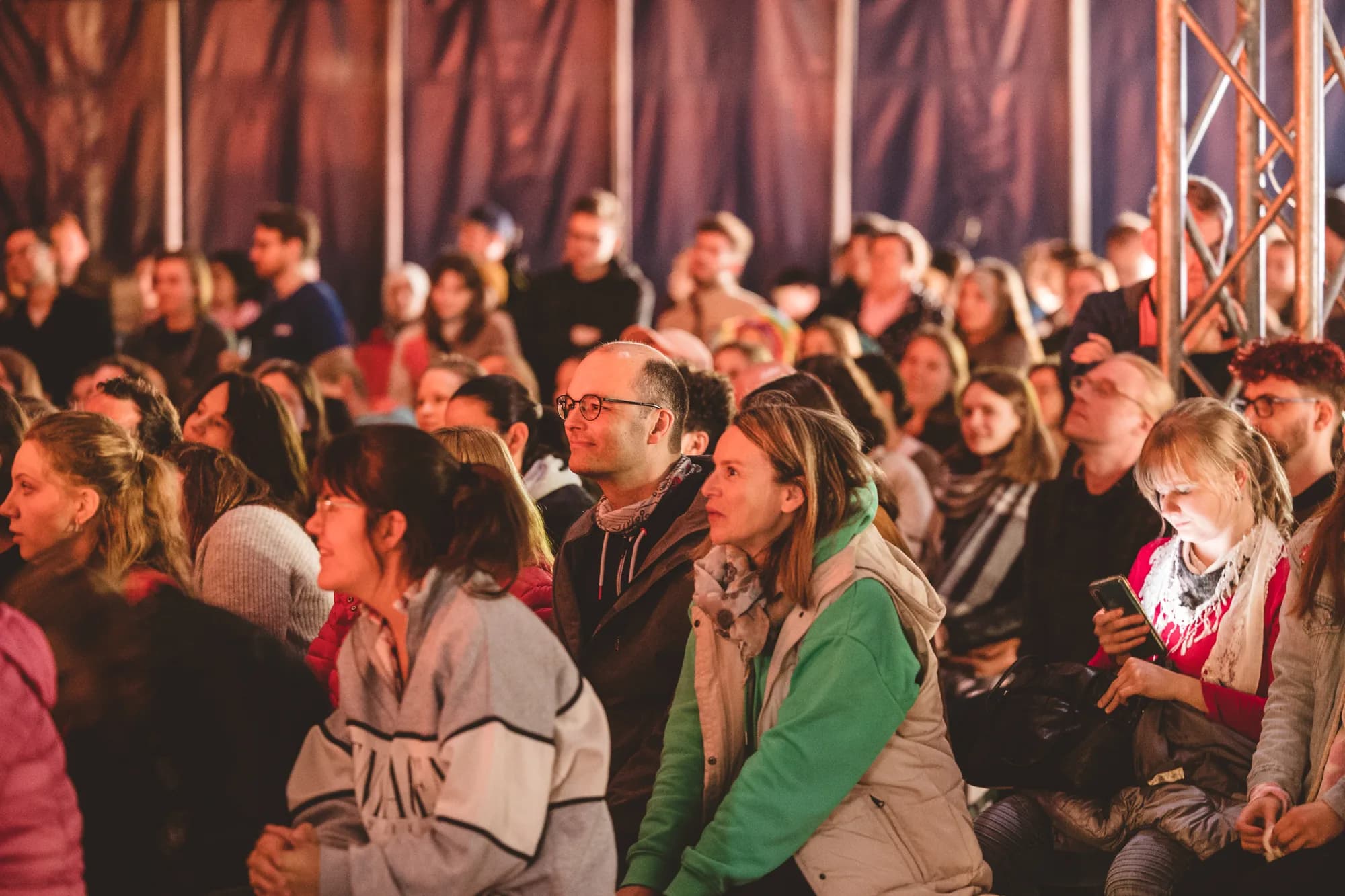 A seated audience of diverse adults watches a performance intently inside a tent with warm lighting.