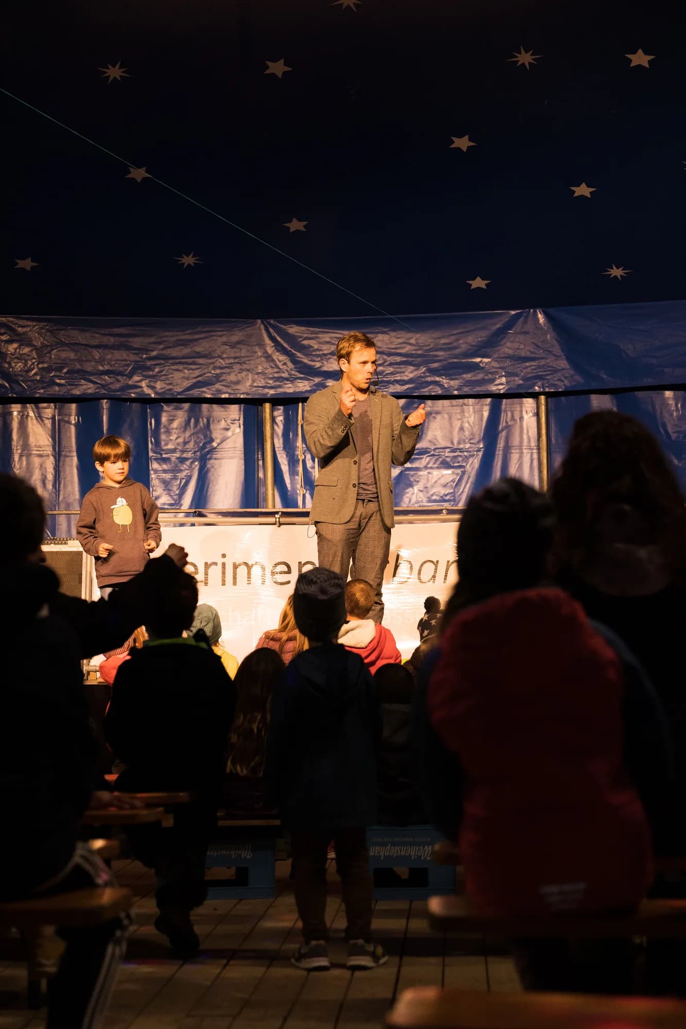 A performer and a young boy stand on a wooden stage under a starry blue tent while an audience watches.