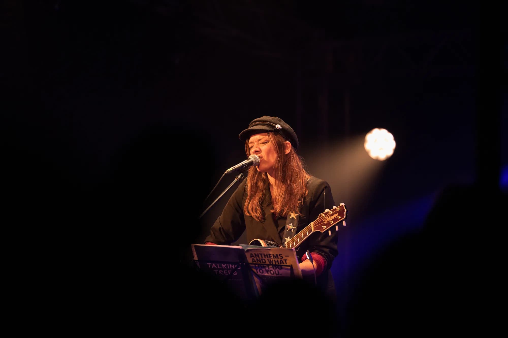 A female musician wearing a cap sings into a microphone while playing an acoustic guitar on a dimly lit stage.