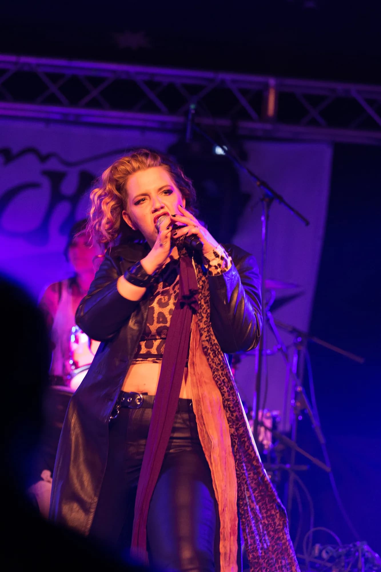 A female rock singer with blonde curly hair performs passionately into a microphone under purple stage lighting.