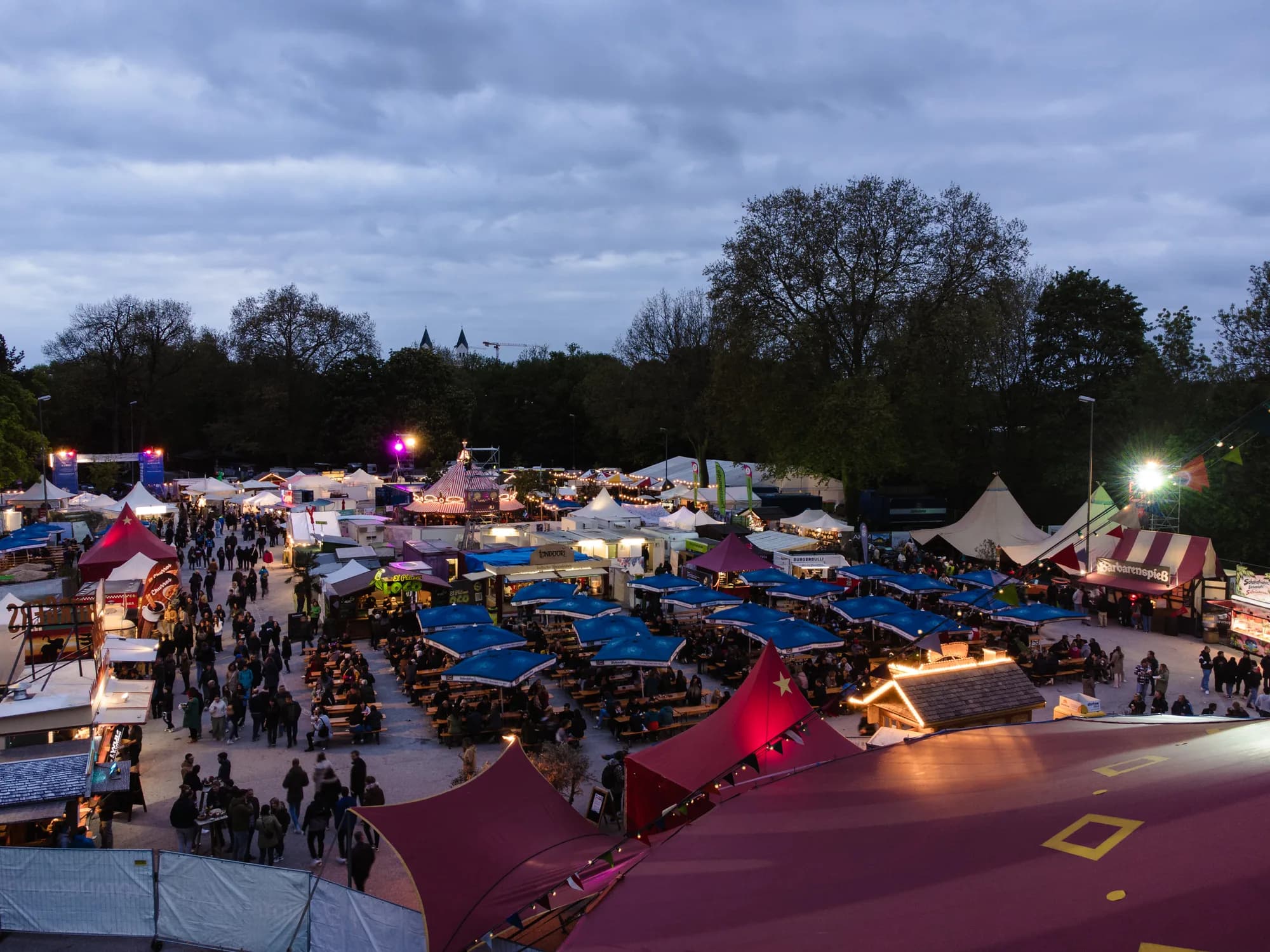 A high-angle drone shot of the Uferlos festival grounds at dusk, showing crowds walking between market stalls and illuminated tents.