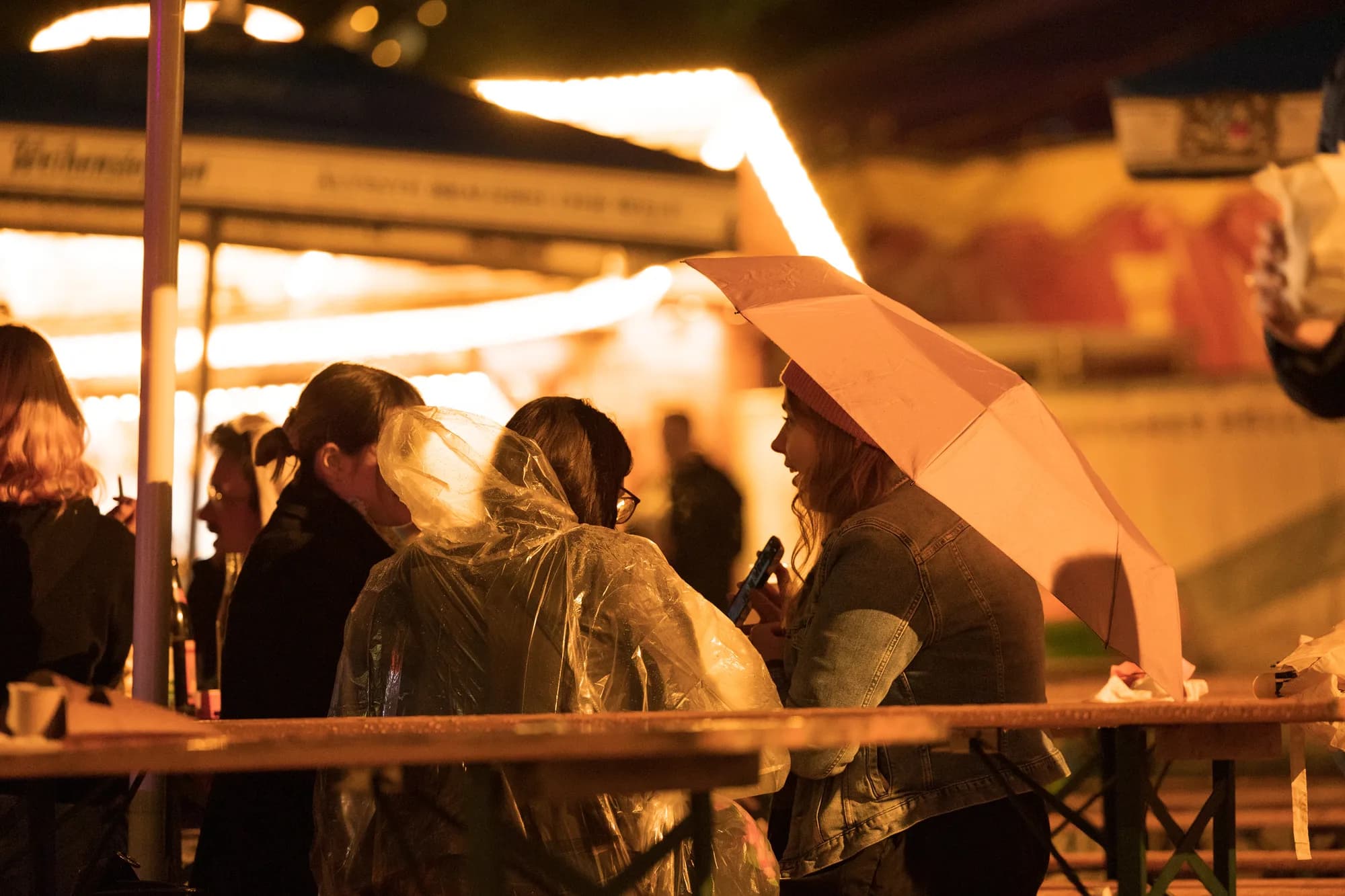 Festival attendees huddle under a pink umbrella and clear plastic ponchos at a wooden table during a rainy evening.