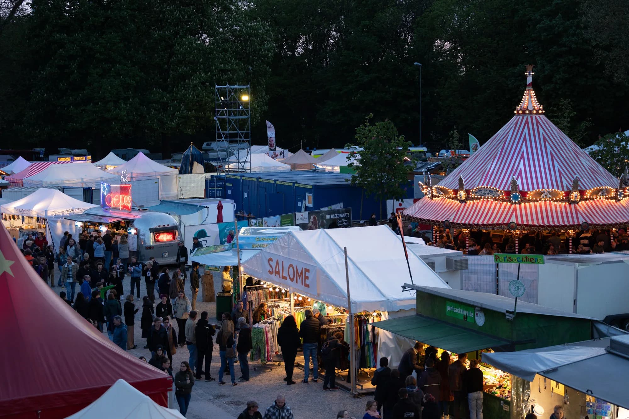 A view of the Uferlos festival market at dusk featuring a lit carousel and white vendor tents.