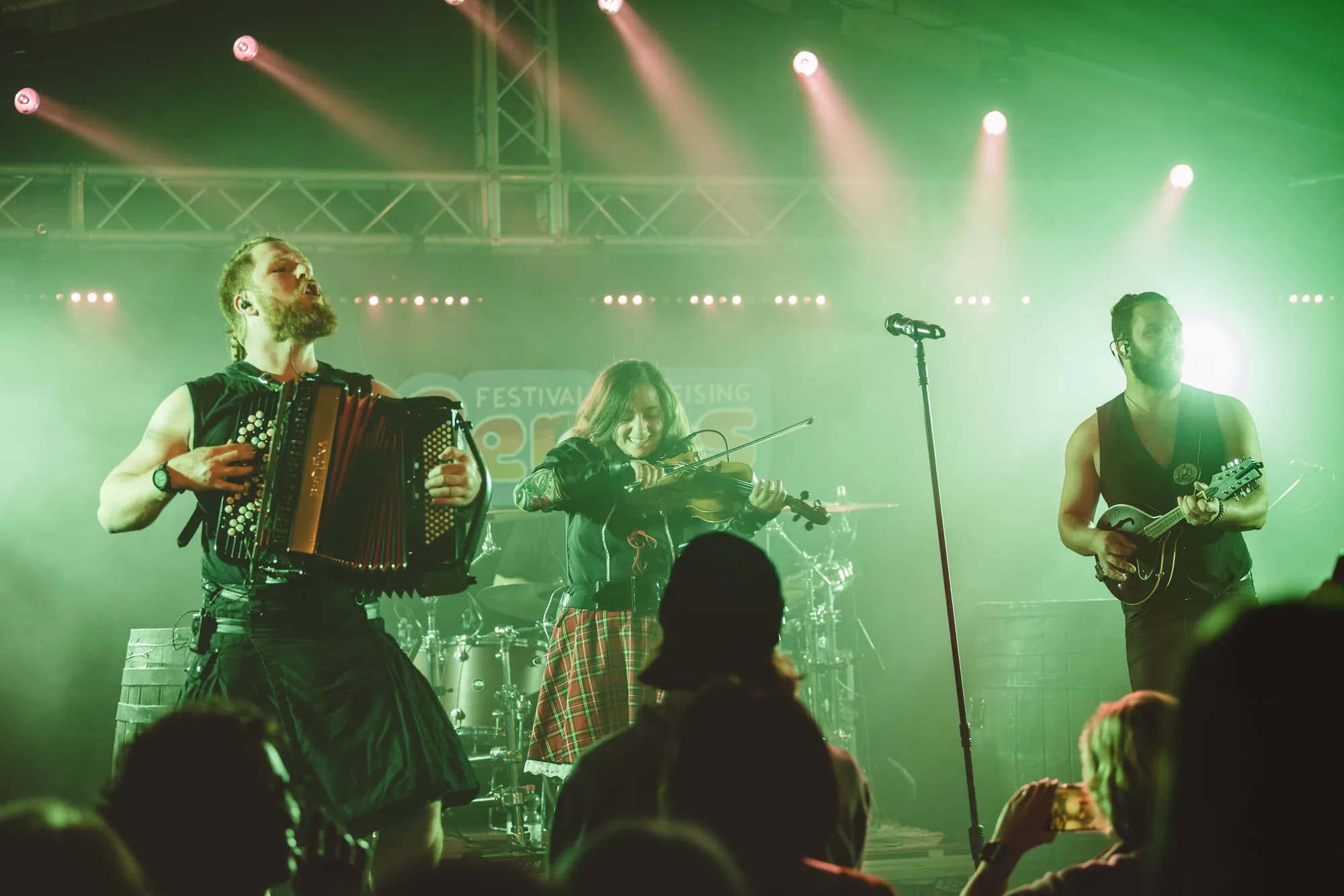 Five musicians perform an acoustic set on stage illuminated by green and orange spotlights while a crowd watches.