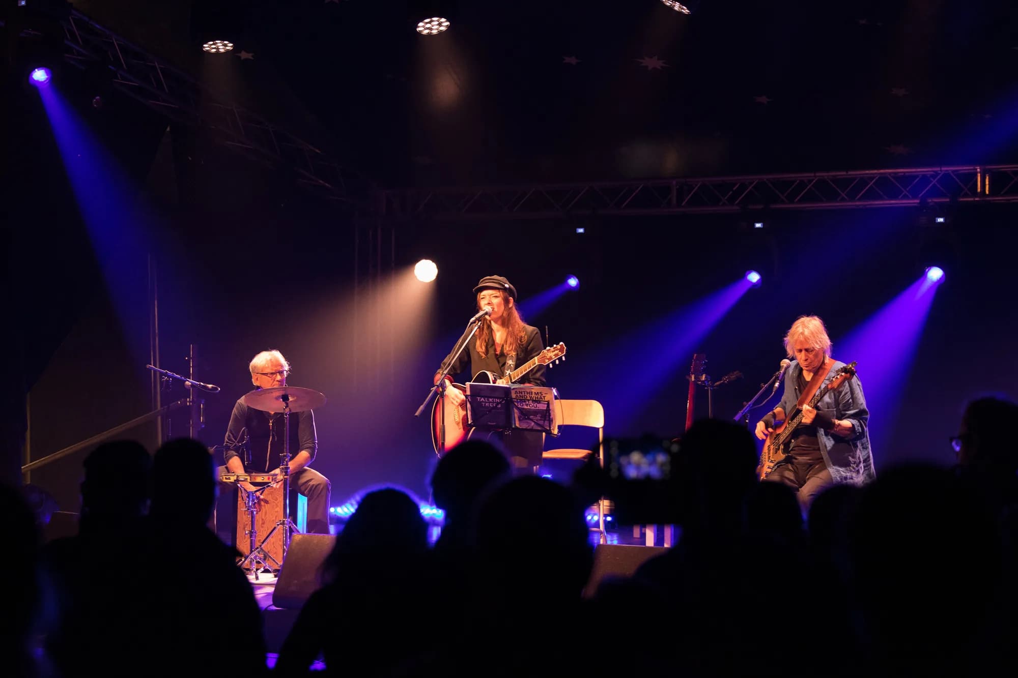 A three-piece band performs on a dimly lit stage illuminated by blue spotlights, featuring a central singer with a guitar and hat, a percussionist on the left, and a guitarist on the right.