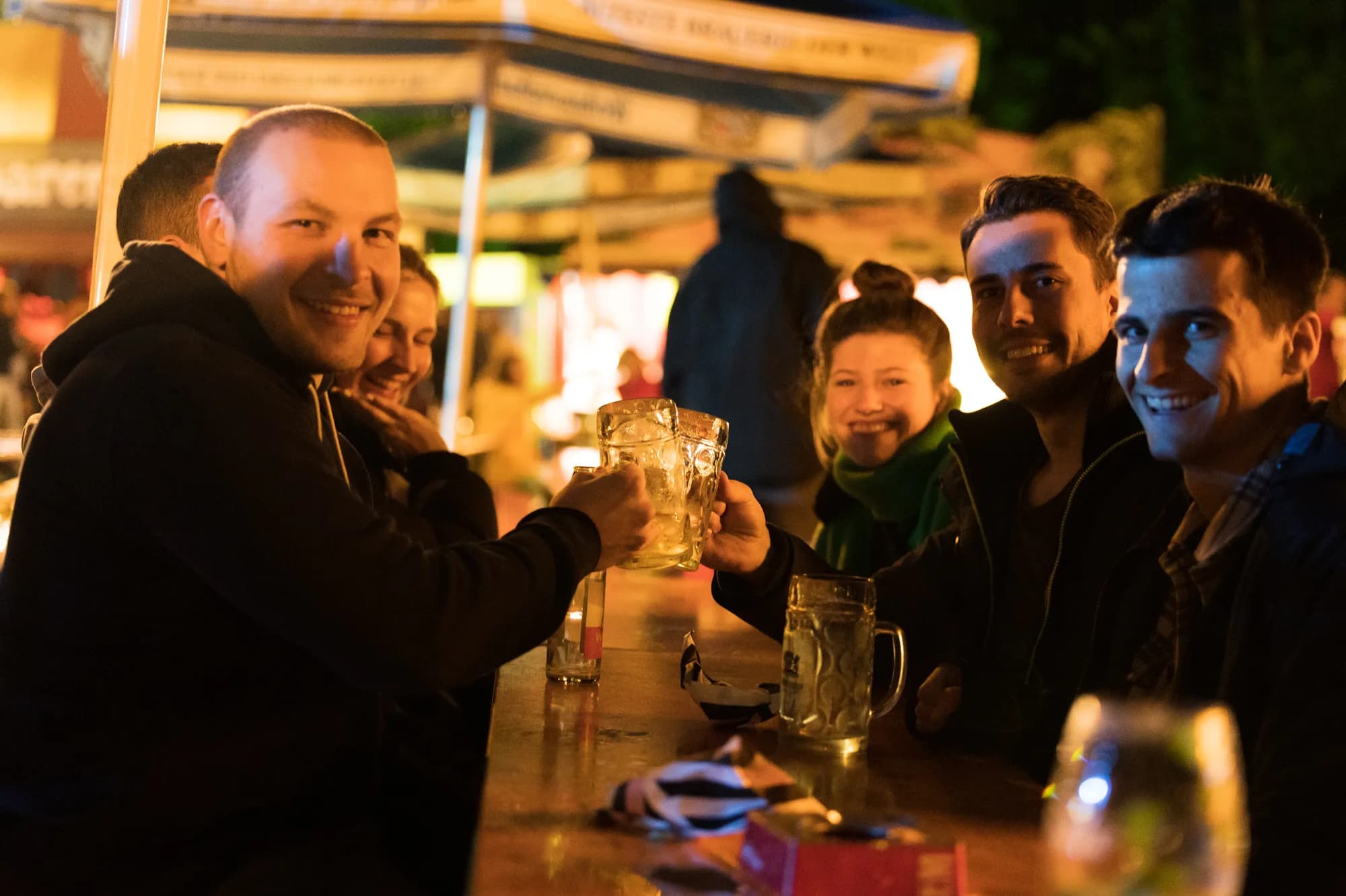 A group of smiling friends clinking beer mugs together at a wooden table in the festival beer garden at night.