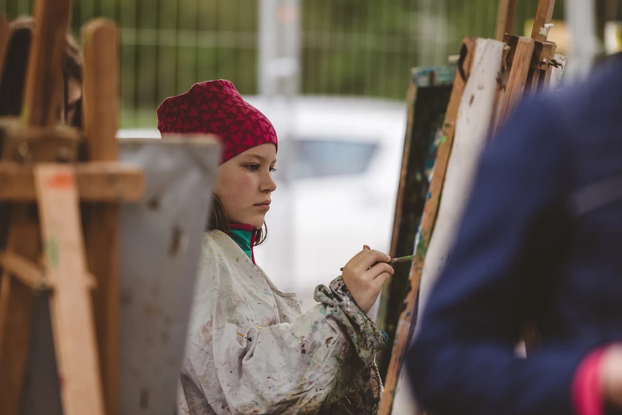 A young girl wearing a red hat and paint-splattered smock concentrates on painting at an easel.