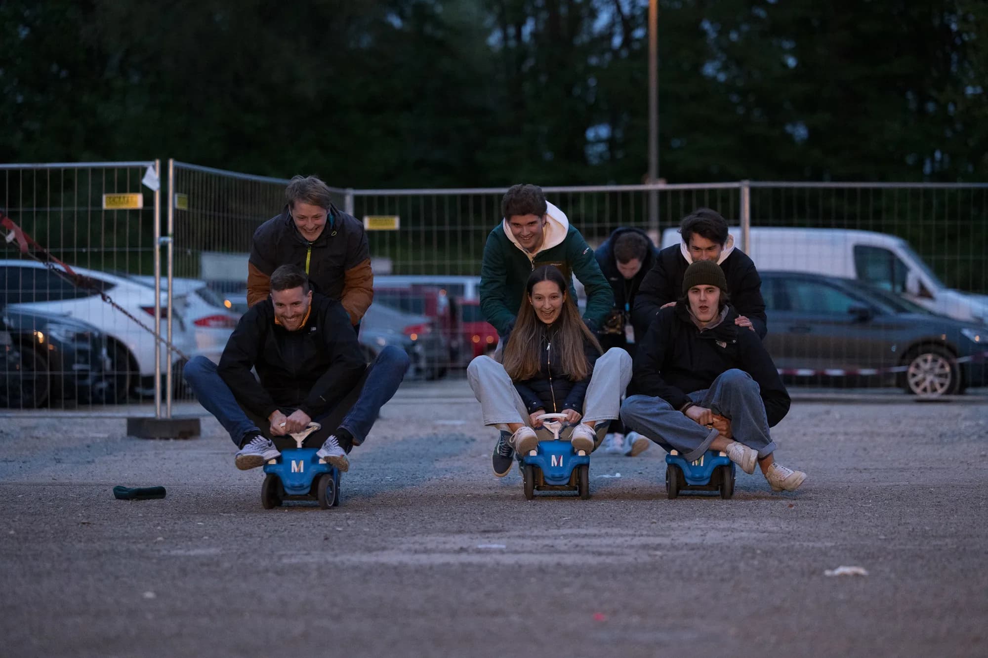 A group of young adults racing on small blue pedal cars in a parking lot at dusk.