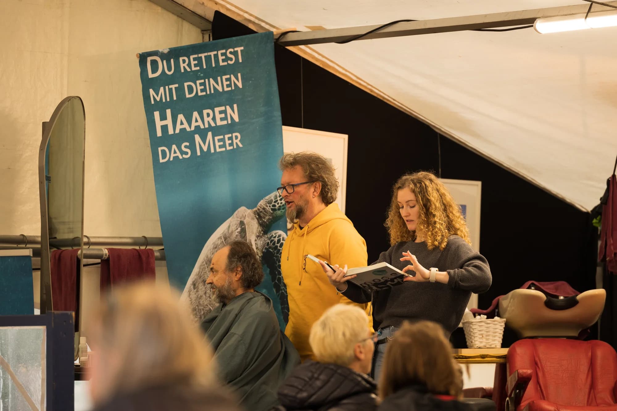A man and woman demonstrate a sustainable hair styling workshop with a banner reading 'Du rettest mit deinen Haaren das Meer' behind them.