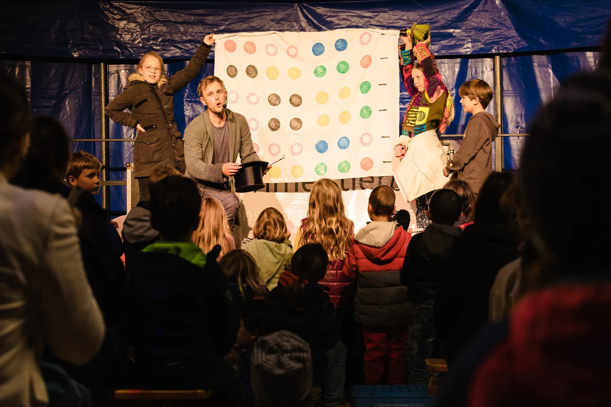 A magician performs a magic trick with a large colorful dot board while children watch from the front row.