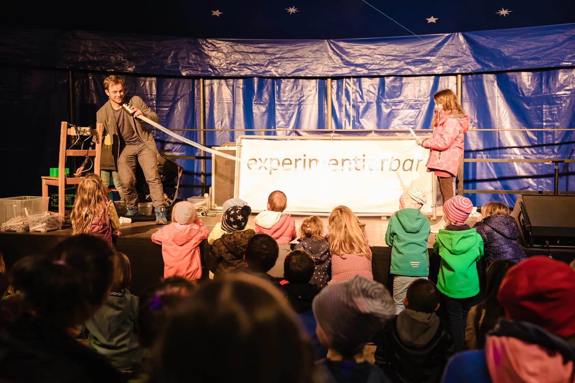 A performer demonstrates a science experiment on stage while a group of children watch attentively.