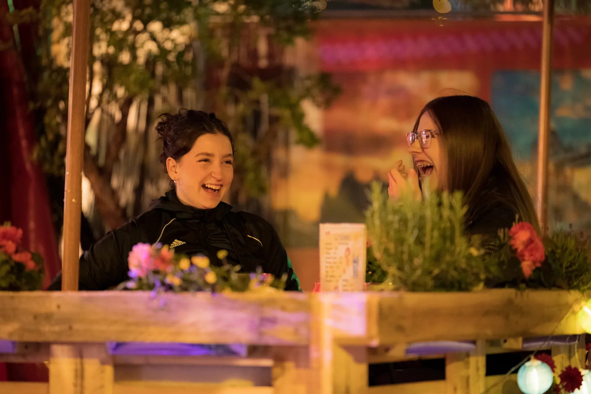 Two young women laughing together while sitting at a wooden table decorated with flowers at night.