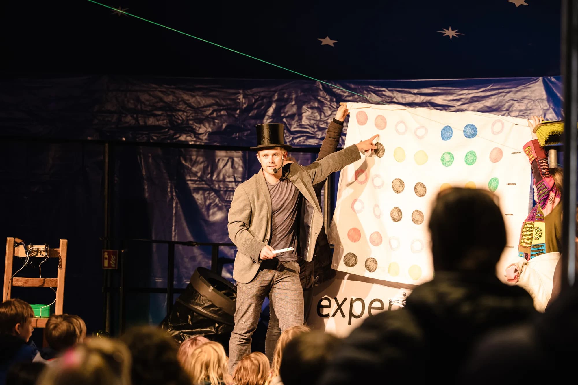 A magician in a top hat performs a trick with a colorful spotted board for an audience of children.