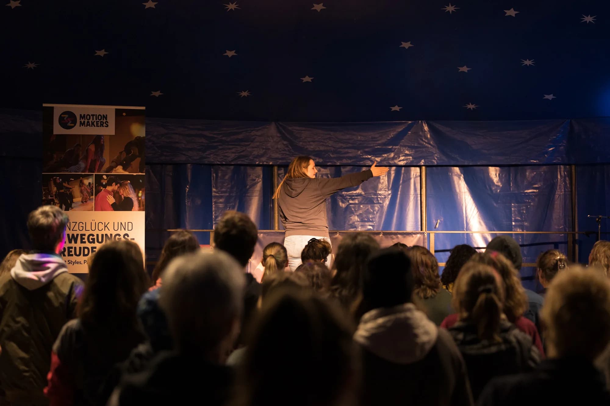 A female instructor on stage gestures to a seated audience during a workshop inside a dark tent with a starry ceiling.