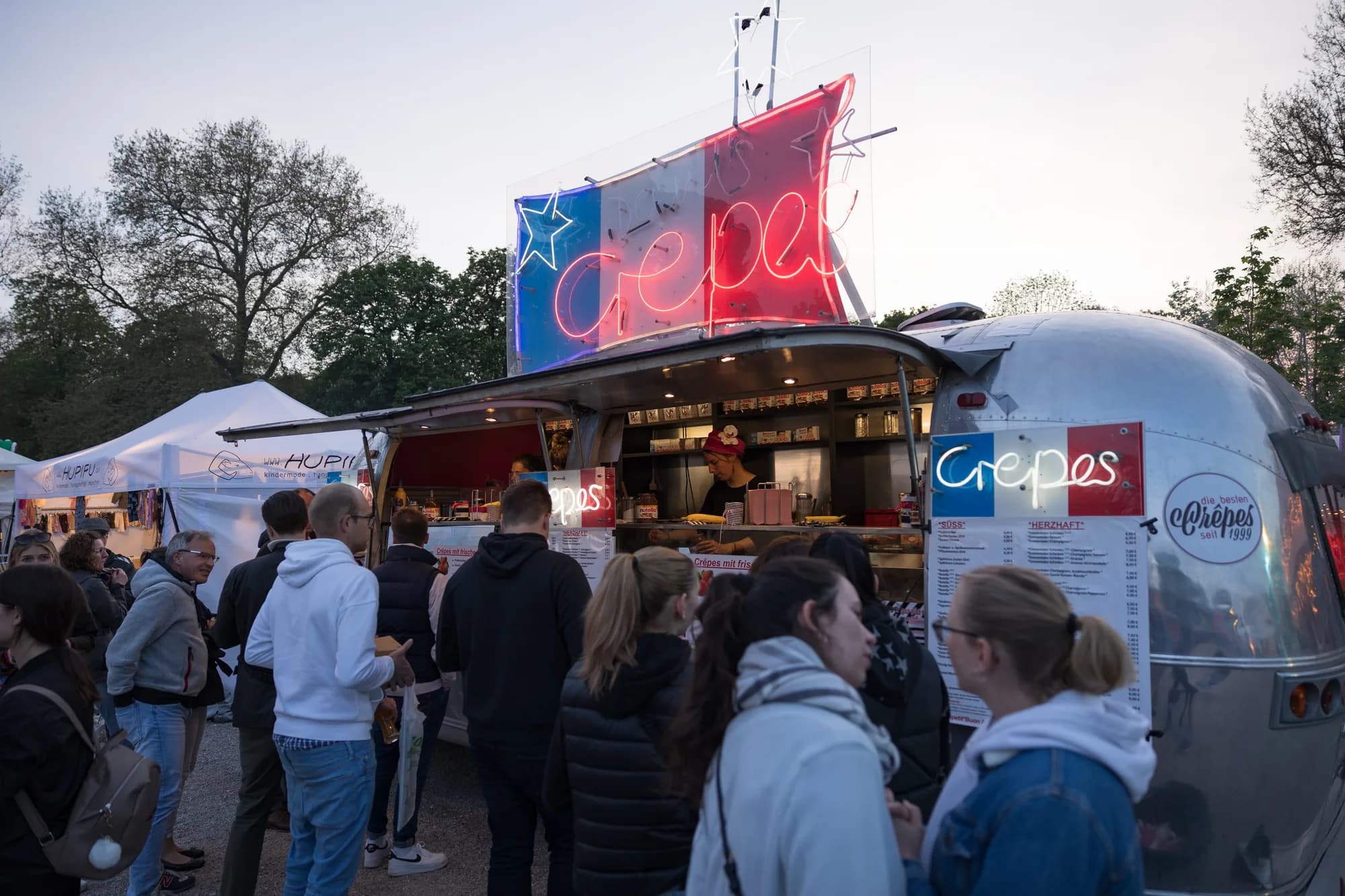 A silver Airstream trailer with a red and blue neon sign reading 'crepes' serves a line of customers at dusk.