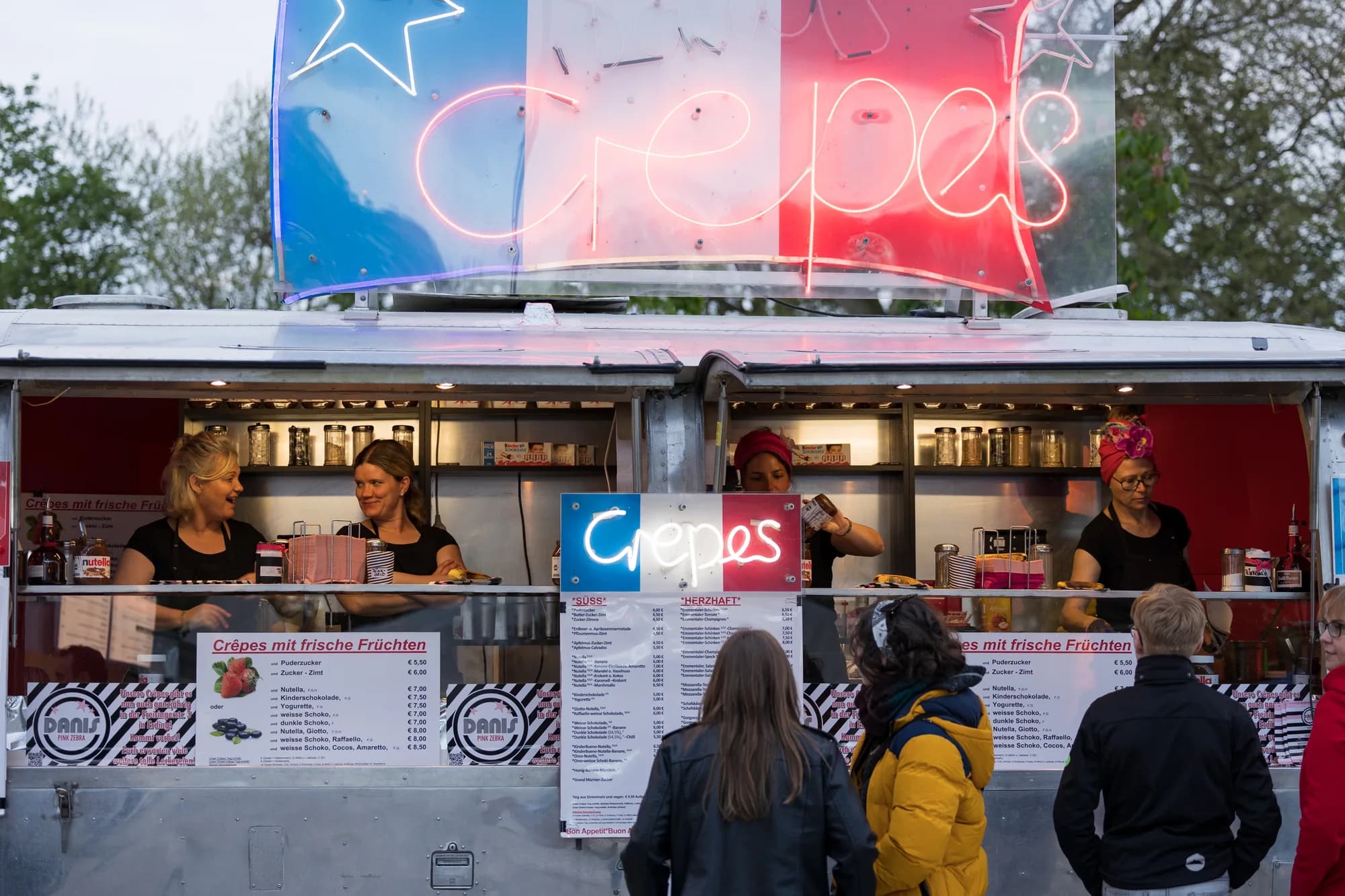 A food truck with a large neon sign selling crepes, with staff serving customers in the evening.
