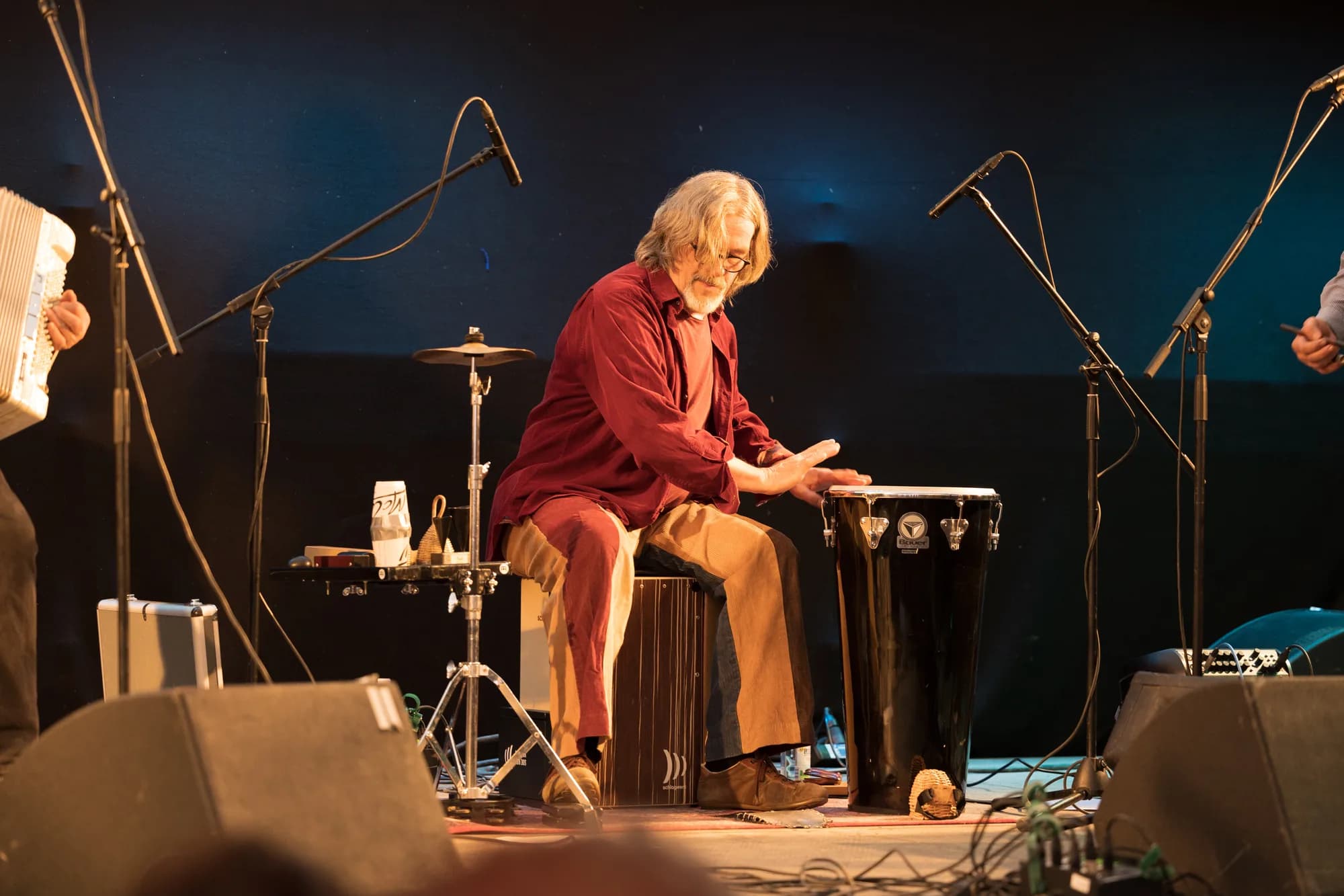 A musician with grey hair and a beard plays a black conga drum while seated on a cajon on a dimly lit stage.