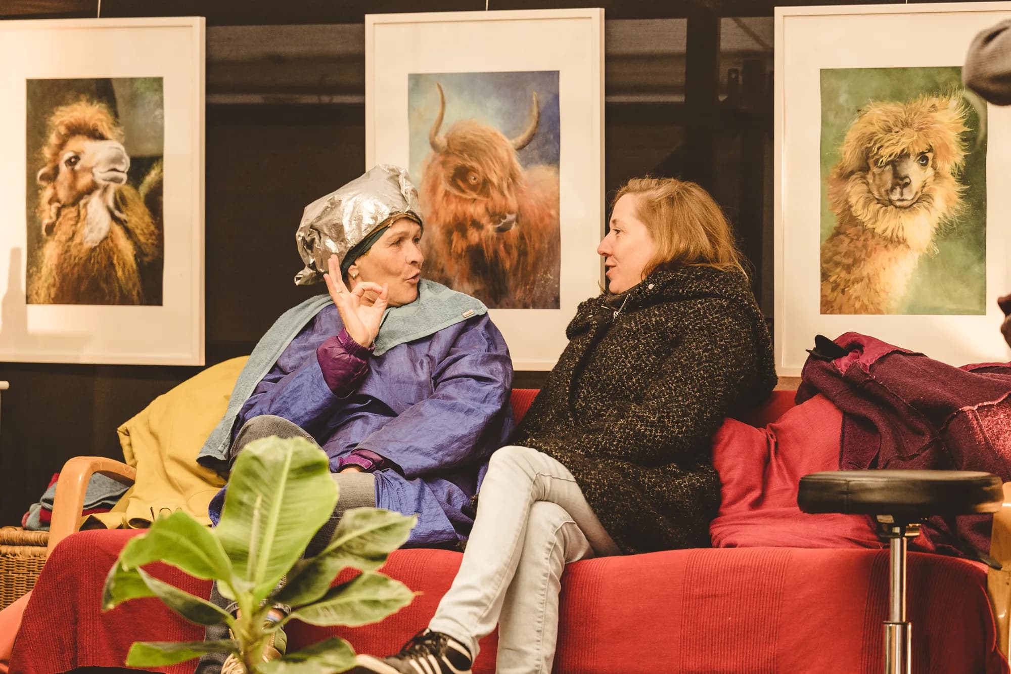 Two performers sit on a red sofa in conversation, one wearing a foil hat, with three large framed animal portraits hanging on the wall behind them.