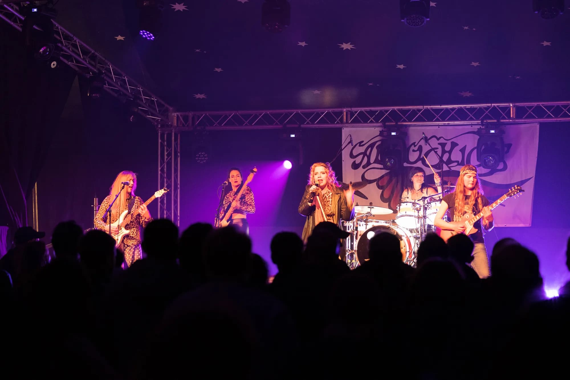 A rock band performs on a stage bathed in purple light with a starry ceiling, playing to a silhouetted crowd.
