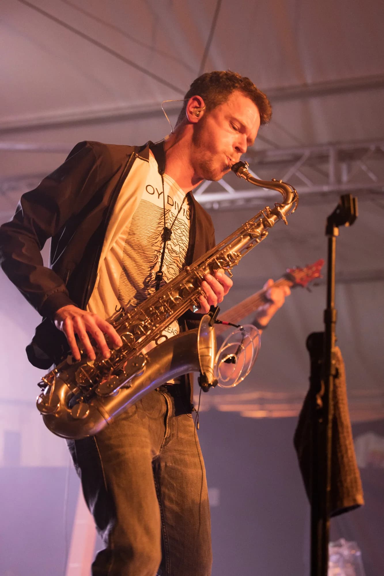 A male musician plays a saxophone on stage wearing a black jacket and white graphic t-shirt.