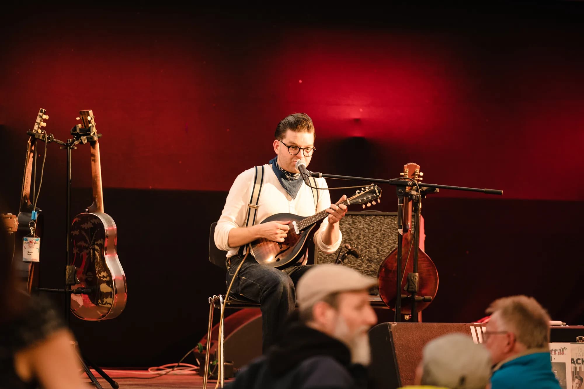 A musician wearing glasses and suspenders plays a mandolin and sings into a microphone on a stage with red lighting.