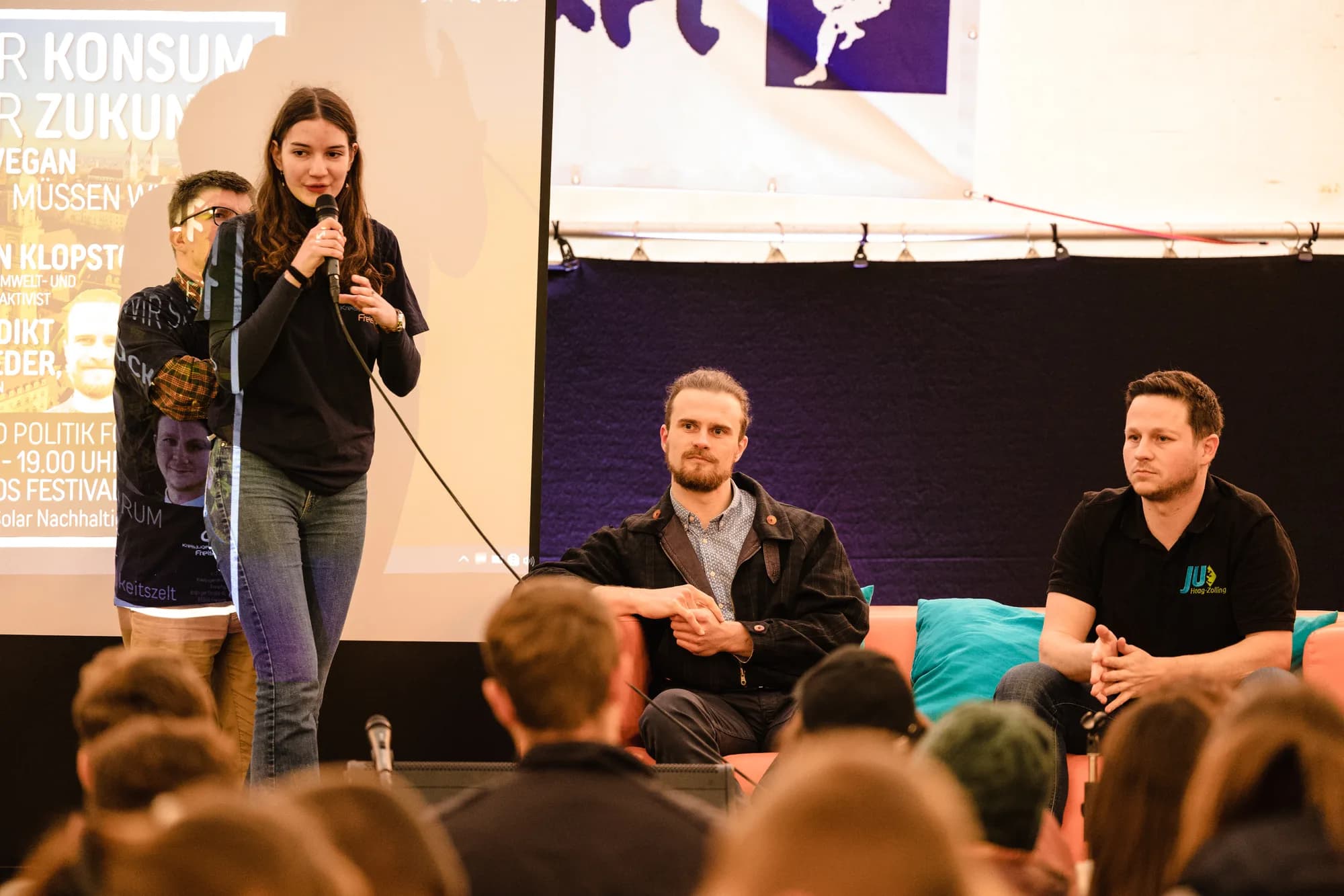 A young woman speaks into a microphone while two men listen during a panel discussion on sustainable consumption.