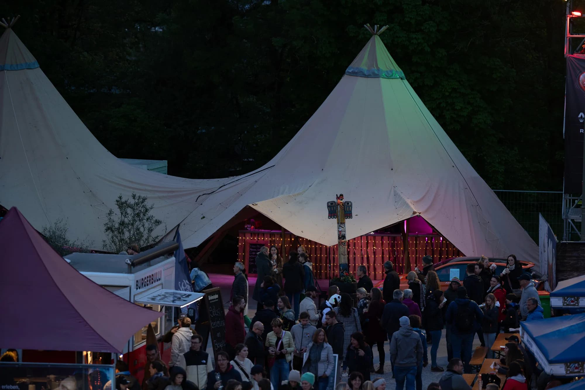A large white tipi tent illuminated with pink and green lights at night, surrounded by a crowd of festival-goers and market stalls.