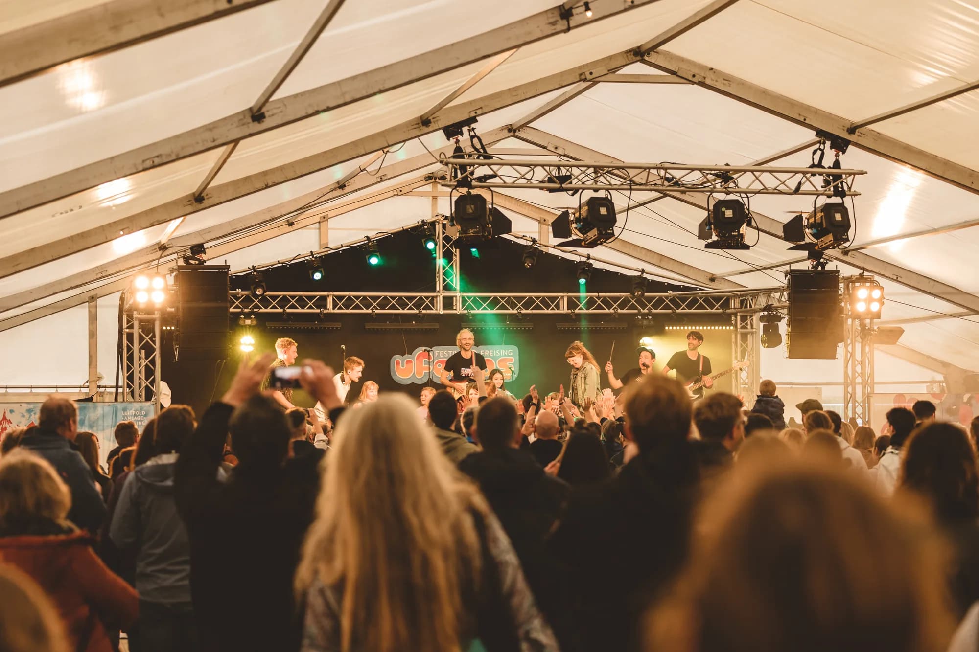 A band performs on stage under a large white tent while the audience watches and records with phones.