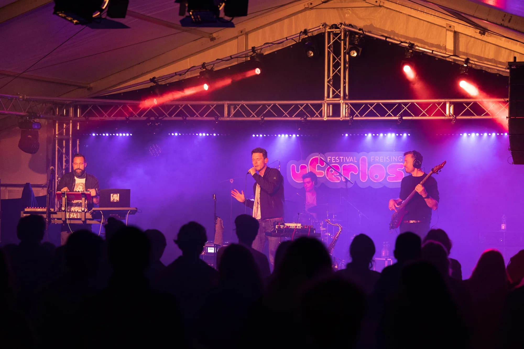 A four-piece band performs on a stage bathed in purple and red light with a crowd watching in silhouette.