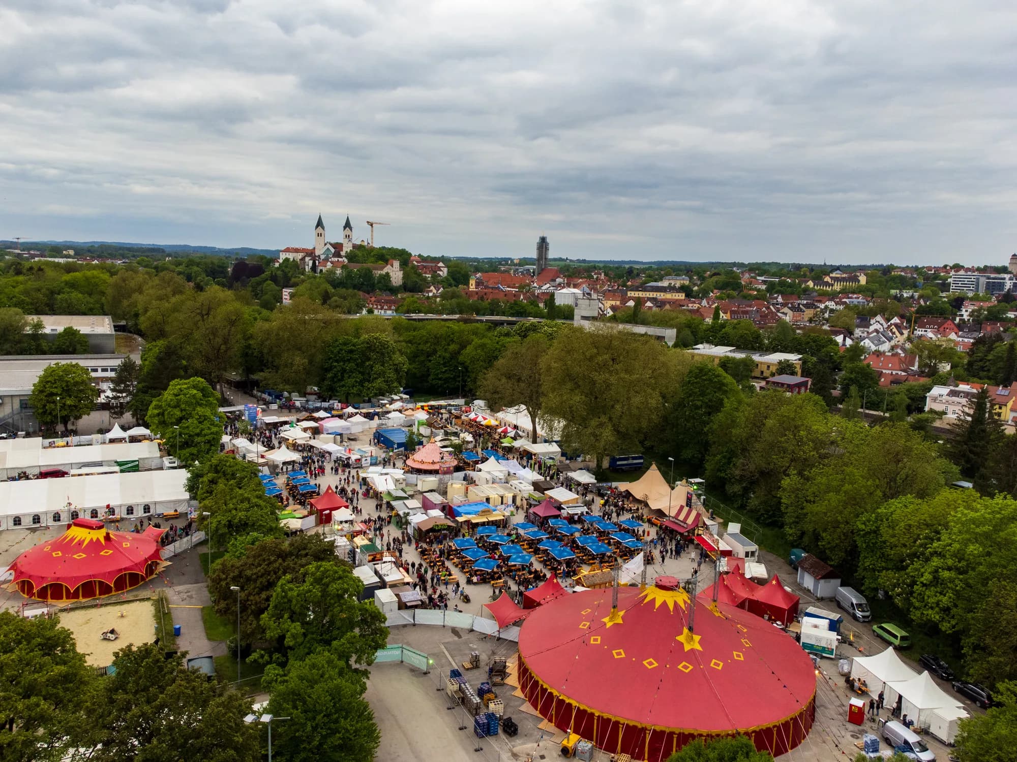 High-angle drone shot of the Uferlos festival grounds with red circus tents and crowds near the Isar river.