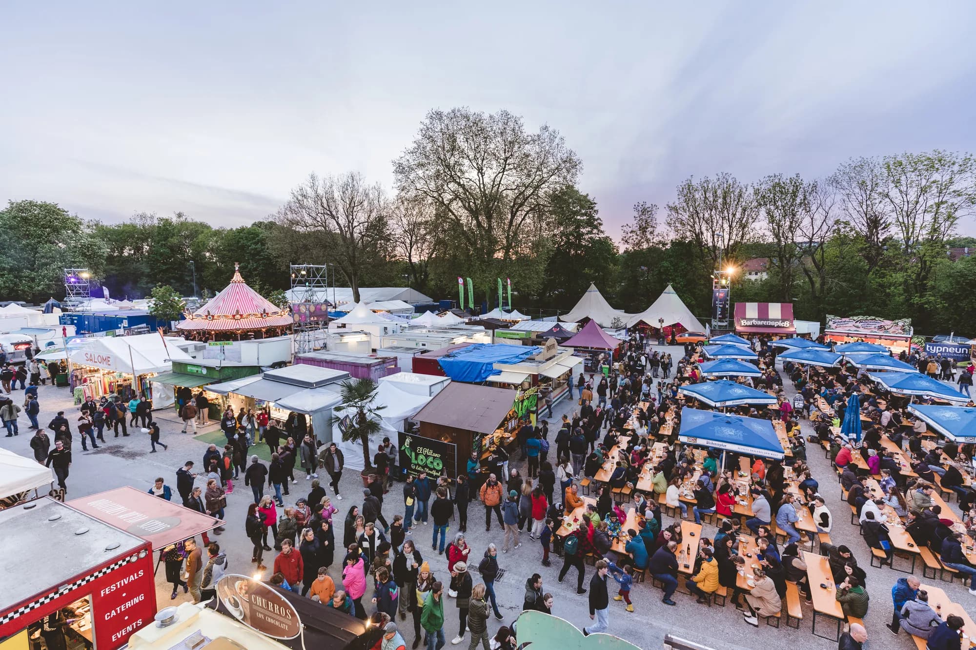 A high-angle drone shot of the Uferlos festival grounds at dusk showing crowds in the beer garden and market stalls.