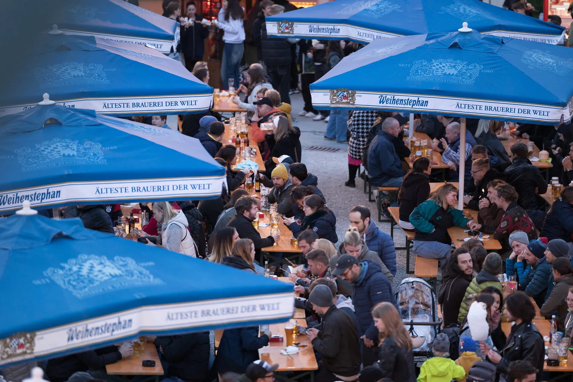 A crowded beer garden scene with guests sitting at long wooden tables under large blue Weihenstephan umbrellas.
