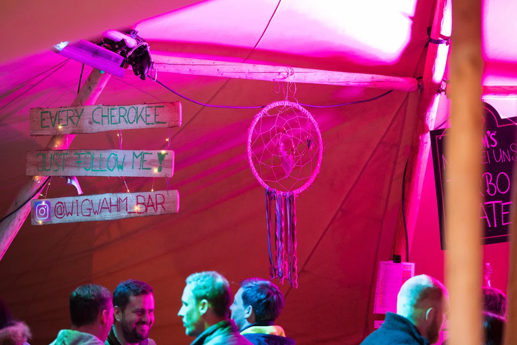 Hand-painted wooden signs reading 'Every Cherokee Just Follow Me' and '@WIGWAHM_BAR' hang inside a tent illuminated by bright pink light.